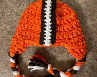 May include: Orange, brown, and white crocheted hat with a long braided strap and tassels. The hat is designed to look like a football helmet.