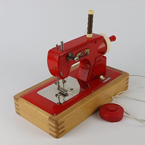 Peut inclure: Une machine à coudre Regina vintage dotée d'un corps en métal rouge vif monté sur une base en bois clair. La machine comprend une plaque à aiguille argentée, un porte-bobine vertical avec du fil marron, un volant et une pédale ronde rouge.