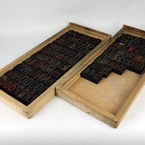 Könnte beinhalten: Zwei offene Holzkisten gefüllt mit alten Letterpress-Druckblöcken. Die Blöcke sind schwarz mit roter und grüner Tinte, in Reihen angeordnet. Die Kisten sind hellbraun und stehen auf einer weißen Oberfläche.