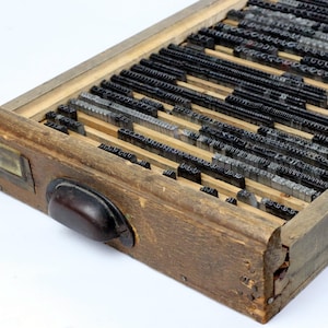 May include: A vintage wooden printer's type case filled with metal letterpress type. The case is rectangular with a dark wooden handle and a metal label plate. The type is arranged in rows, showing various fonts and sizes, ideal for printing.