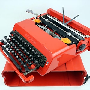 Peut inclure: Une machine à écrire vintage rouge vif avec des touches noires et le mot "valentine" imprimé sur le devant. La machine à écrire est ouverte, révélant les mécanismes internes, et repose sur son étui de transport rouge assorti.