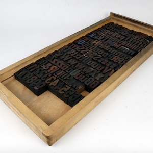 Könnte beinhalten: Eine hölzerne Druckertablett mit einzelnen Letterpress-Druckblöcken. Die Blöcke sind hauptsächlich schwarz mit einigen Buchstaben in Rot und Blau. Das Tablett ist rechteckig und aus hellem Holz gefertigt und zeigt eine Vintage-Ästhetik.