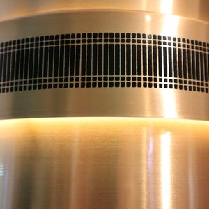 May include: A copper-colored metal lamp with a black geometric pattern around the middle. The lamp has a white light shining from the top.