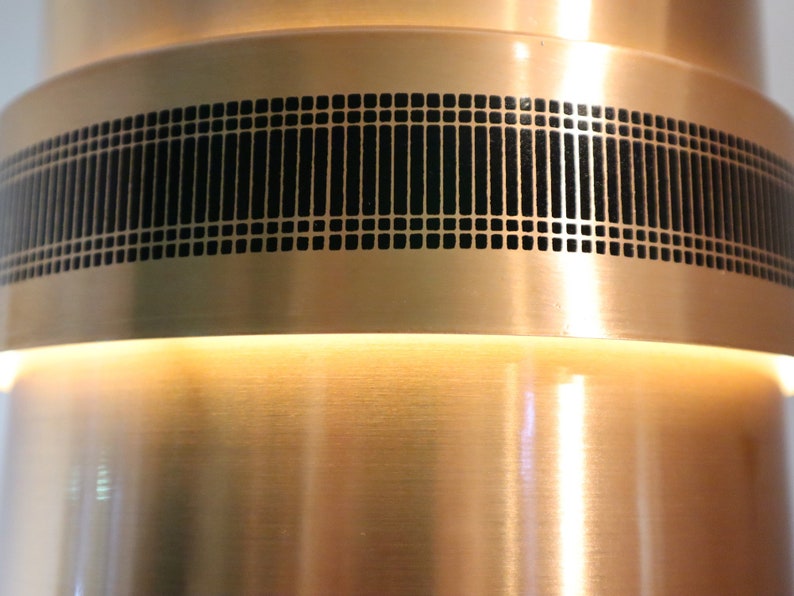 May include: A copper-colored metal lamp with a black geometric pattern around the middle. The lamp has a white light shining from the top.