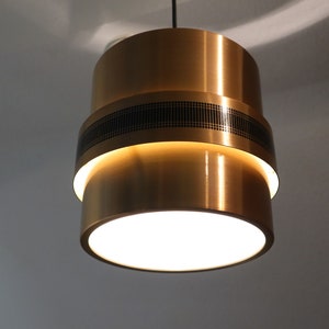 May include: A gold-toned pendant light with a black geometric pattern on the middle band. The light is on and illuminating the room.