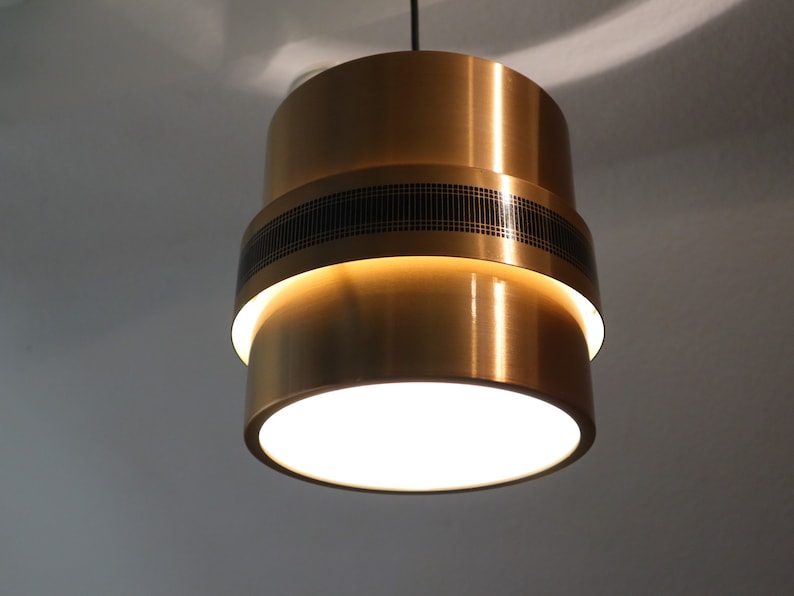 May include: A gold-toned pendant light with a black geometric pattern on the middle band. The light is on and illuminating the room.