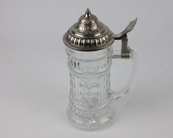 May include: A clear glass beer stein with a silver lid and handle. The stein has a decorative pattern on the body.
