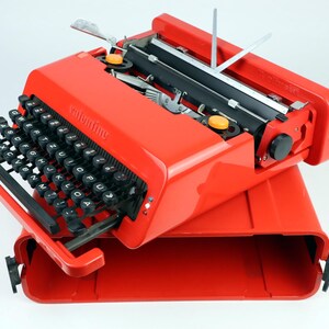 May include: A bright red portable typewriter with black keys and the word valentine embossed on the front casing. The typewriter features a black carriage, two yellow circular knobs, and a matching red carrying case base underneath the unit.