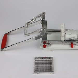 May include: A silver-colored, manual French fry cutter with a red handle. The cutter sits on a white base and includes a separate cutting grid. The device is designed to slice potatoes into uniform strips for frying.