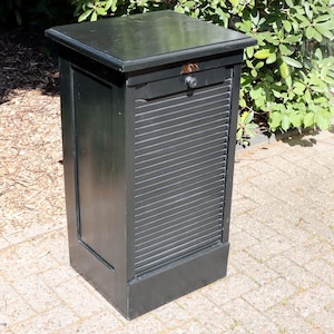 Könnte beinhalten: Ein schwarzer Holzschrank mit flacher Oberseite und einer Lamellenfront. Der Schrank hat eine kleine Schublade mit einem runden Knopf. Der Schrank ist rechteckig und steht auf einer gepflasterten Oberfläche.