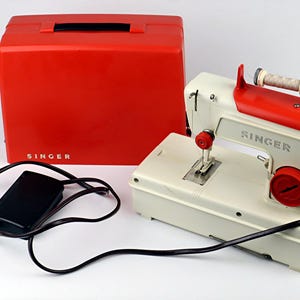 Könnte beinhalten: Eine cremefarbene Singer-Nähmaschine mit roten Akzenten, einem roten Tragekoffer mit Singer-Logo und einem schwarzen Fußpedal mit Stromkabel. Die Maschine verfügt über einen oben angebrachten Garnrollenhalter und ein rotes Einstellrad.
