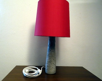 May include: A table lamp featuring a vibrant red cylindrical lampshade paired with a textured, tapered base that transitions from a dark gray at the top to a lighter, mottled gray at the bottom, complete with a white electrical cord and plug.
