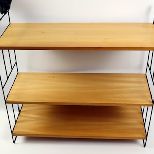 May include: A three-tiered wooden shelf unit with a black metal frame. The shelves are made of light wood and the frame is made of thin black metal.