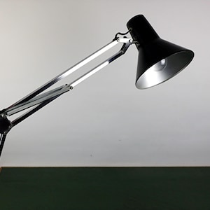 May include: A black desk lamp with a silver interior shade. The lamp has a long, adjustable arm with a spring mechanism and a black base. The lamp is angled towards the viewer, with the light source visible.
