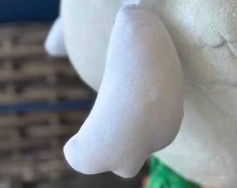 May include: A white fabric wing attached to a white body with a thin white ribbon strap. The wing has a soft, rounded shape and is positioned against a textured white surface, with a glimpse of green and orange elements visible at the base.