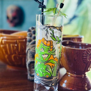 May include: A tall, clear glass with a green and orange cartoon creature design. The glass has a black straw and a small green paper umbrella. Other tiki-themed serving ware is in the background.