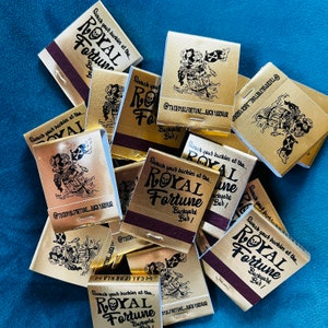 May include: A collection of gold matchbooks with a maroon striking strip. Each matchbook features the text "ROYAL FORTUNE Backyard Bar!" and a pirate-themed illustration. The text "Swash your buckles at the..." is also present.