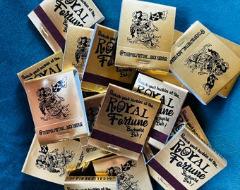 May include: A collection of gold matchbooks with a maroon striking strip. Each matchbook features the text "ROYAL FORTUNE Backyard Bar!" and a pirate-themed illustration. The text "Swash your buckles at the..." is also present.