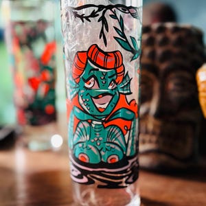 May include: A clear glass tumbler with a colorful tiki-style design. The illustration features a teal-skinned creature with orange accents, set against a black and white water-like pattern. The glass is filled with a clear material.
