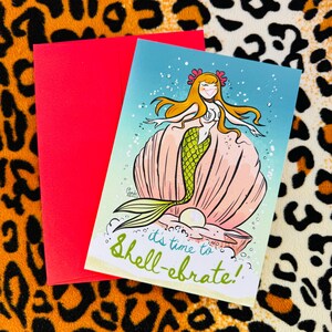 May include: A greeting card featuring a cartoon mermaid in a seashell. The mermaid has long blonde hair and is wearing a green tail. The card reads "It's time to shell-ebrate!"