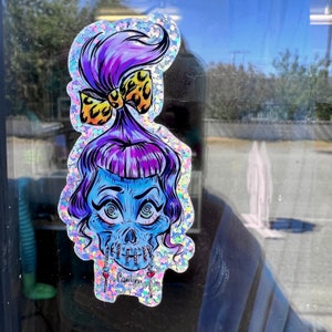May include: A holographic sticker featuring a stylized skull with blue skin, purple hair, and a leopard print bow. The skull has spiral eyes and dangling heart-shaped earrings. The sticker is outlined with iridescent glitter.