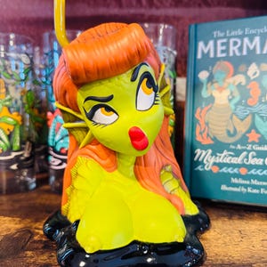 May include: A ceramic mermaid tiki mug with a yellow-green face, orange hair, and a red lip. The mug has a black base and a yellow snorkel. A book titled "The Little Encyclopedia of Mermaids" is in the background.