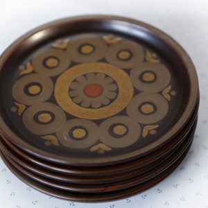 May include: A stack of six brown ceramic plates with a gold and brown geometric pattern. The plates have a raised rim and a smooth surface.