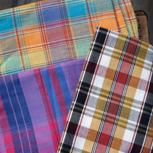 May include: Three pieces of fabric with different plaid patterns. The top fabric is a light orange, yellow, and blue plaid. The middle fabric is a purple and blue plaid. The bottom fabric is a black, white, red, and yellow plaid.
