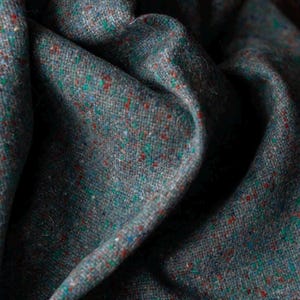 Może przedstawiać: Zbliżenie na teksturowaną tkaninę z szarą bazą, nakrapianą czerwonymi, zielonymi i niebieskimi plamkami. Tkanina jest udrapowana, ukazując fałdy i cienie. Materiał wydaje się być tkanym tweedem, odpowiednim do odzieży lub wystroju wnętrz.