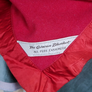 May include: Close-up of a red cashmere blanket with a satin border. The blanket has a label that reads "The Carwan Blanket, All Pure Cashmere". The blanket is folded, showing the texture and color.