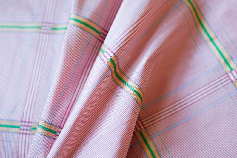 May include: Pink and white checkered fabric with thin green, yellow, and blue stripes. The fabric is folded and has a soft, textured appearance.