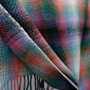 May include: A close-up of a colorful plaid fabric with a fringed edge. The fabric is folded in a way that shows the different colors and patterns.