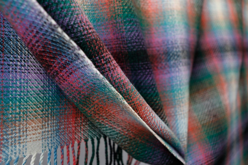 May include: A close-up of a colorful plaid fabric with a fringed edge. The fabric is folded in a way that shows the different colors and patterns.