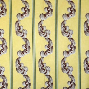 Op de afbeelding: Een gele stof met een herhalend patroon van grijze en bruine gestileerde bladeren. De bladeren zijn in een golvend patroon gerangschikt en worden gescheiden door dunne groene strepen.