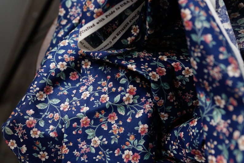Pu&ograve; includere: Primo piano di un tessuto blu navy con un motivo floreale bianco e rosa. Il tessuto &egrave; piegato e ha un'etichetta bianca con il testo "Crafted with Pride in USA".