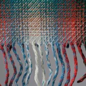 May include: Close-up of a woven fabric with a plaid pattern in shades of red, green, blue, and white. The fabric has a fringe border.