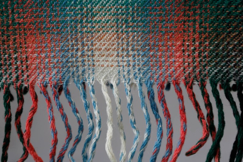 May include: Close-up of a woven fabric with a plaid pattern in shades of red, green, blue, and white. The fabric has a fringe border.