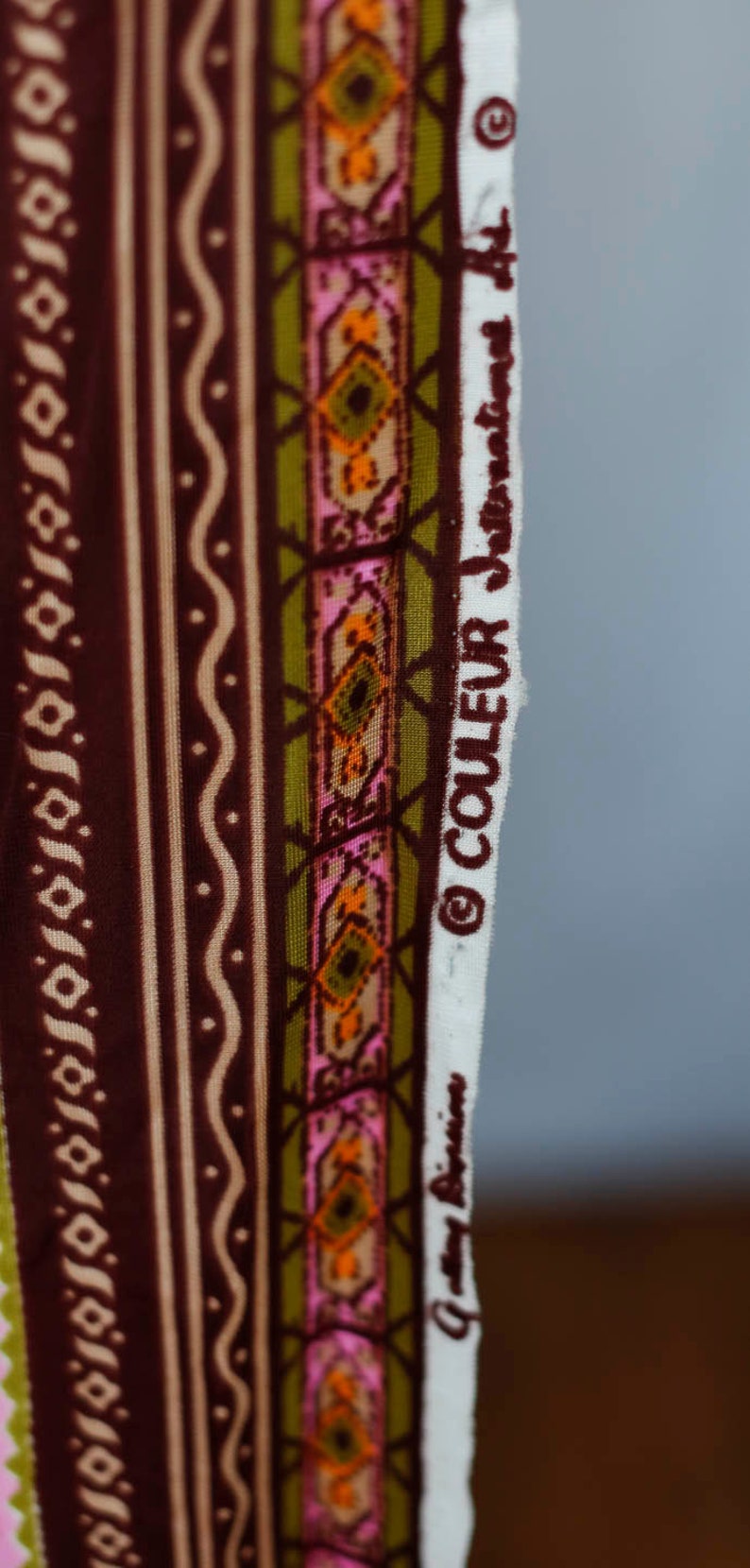 Puede incluir: Primer plano de una tela con un patr&oacute;n geom&eacute;trico marr&oacute;n, verde, rosa y naranja. La tela tiene una etiqueta blanca con el texto "COULEUR" y un s&iacute;mbolo de copyright.