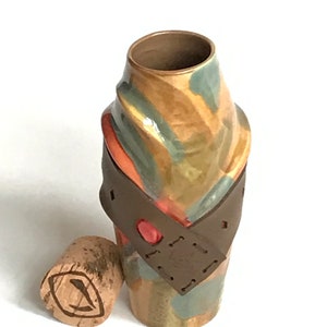 May include: A handcrafted ceramic bottle with a brown leather strap and a cork stopper. The bottle features a unique design with shades of brown, orange, and green. The cork has a painted eye design. A decorative piece for the home.