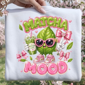May include: A white sweatshirt featuring a graphic of a green leaf character wearing pink sunglasses and pink sneakers. The character holds a green matcha drink with a label reading MATCHA LOVE. The text reads MATCHA MOOD, surrounded by pink bows.