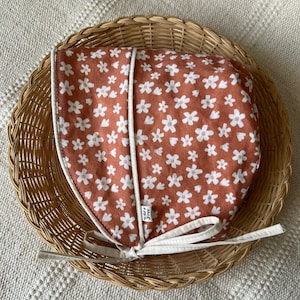 Peut inclure: Un bonnet de couleur terre cuite avec un imprimé floral blanc et une bordure blanche. Le bonnet a un lien à l'arrière. Le bonnet est placé dans un panier en osier.