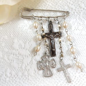 May include: A silver safety pin brooch with multiple silver and brown cross charms and white pearl beads hanging from it.