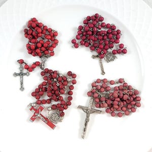 May include: Four rosaries with red wooden beads and silver crucifixes are arranged on a white plate. The beads vary slightly in shade, from a deep red to a lighter, more muted tone. The crucifixes are detailed.