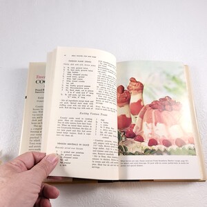 May include: Open vintage cookbook with a recipe for Swedish Frank Steaks and a photo of a strawberry dessert. The cookbook has a light blue cover with the title "Freezing Cookbook" and the words "Prized Recipes of America" printed on the front cover.