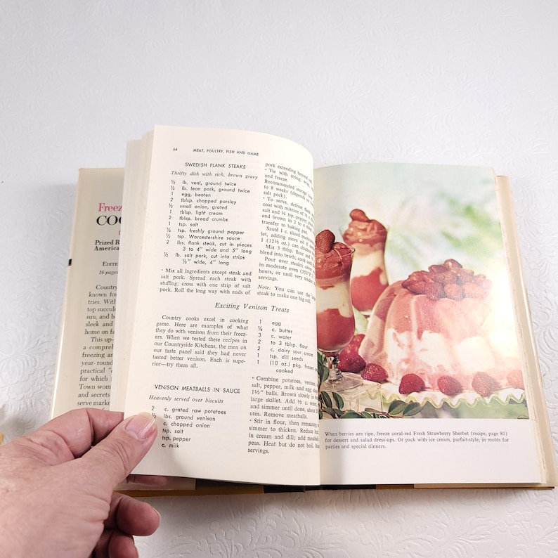 May include: Open vintage cookbook with a recipe for Swedish Frank Steaks and a photo of a strawberry dessert. The cookbook has a light blue cover with the title "Freezing Cookbook" and the words "Prized Recipes of America" printed on the front cover.