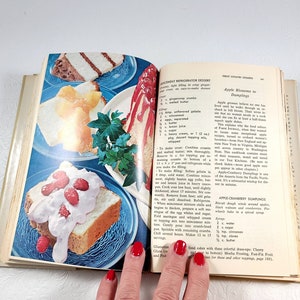 May include: An open cookbook displays colorful images of desserts, including layered cakes with fruit toppings and whipped cream, on blue plates. The pages feature recipes for refrigerator desserts and apple dumplings.