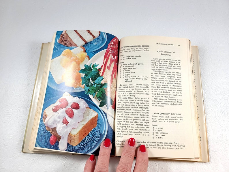 May include: An open cookbook displays colorful images of desserts, including layered cakes with fruit toppings and whipped cream, on blue plates. The pages feature recipes for refrigerator desserts and apple dumplings.