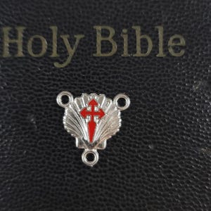 May include: A silver-colored religious charm with a red cross design, set against a black, textured background. The charm is in the shape of a shell. The words "Holy Bible" are visible in gold lettering above the charm.