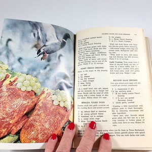 May include: Open cookbook page featuring a vintage illustration of a duck in flight above a salad with grapes and meat. The page includes recipes for salad dressings and salads.