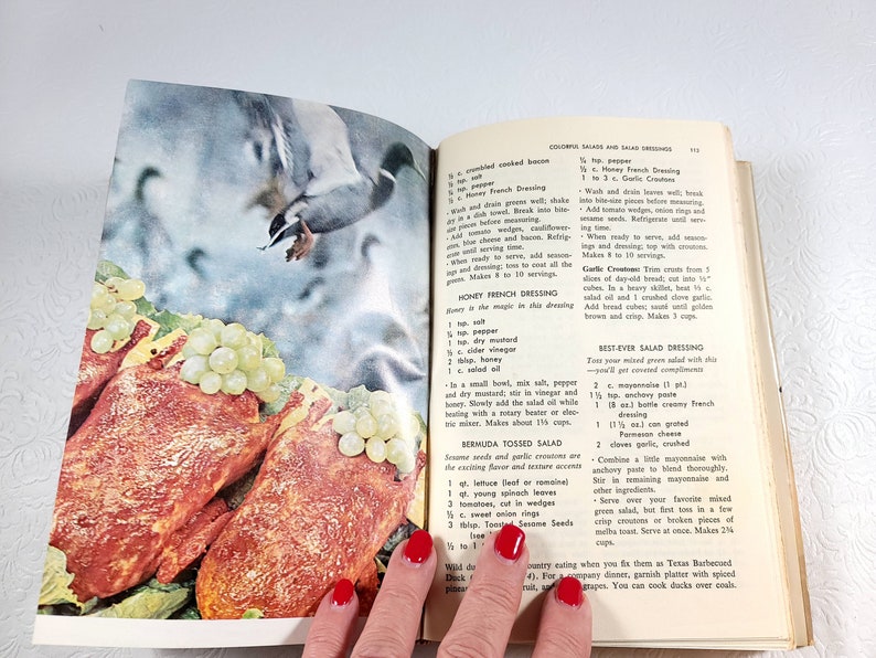 May include: Open cookbook page featuring a vintage illustration of a duck in flight above a salad with grapes and meat. The page includes recipes for salad dressings and salads.